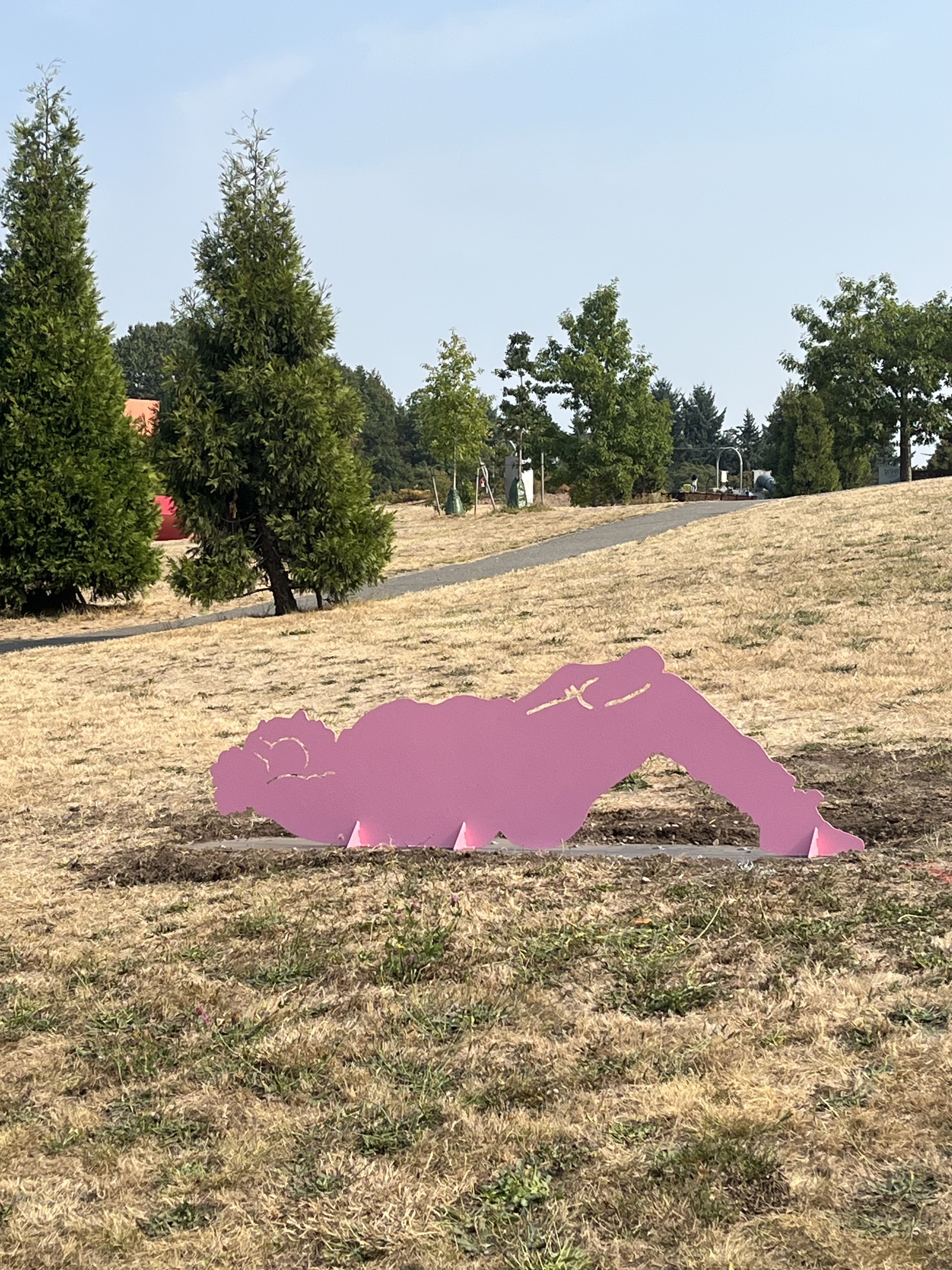 A large pink metal sculpture of a person lying in the grass, relaxing. The setting is a park with trails and trees surrounding.