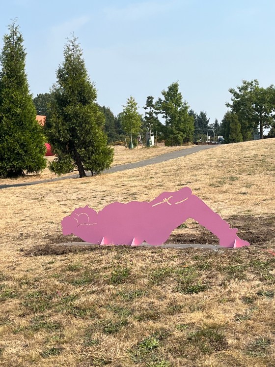 A large pink metal sculpture of a person lying in the grass, relaxing. The setting is a park with trails and trees surrounding.